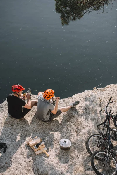 High angle view of bike travellers eating canned food on rocky cliff over lake — Stock Photo