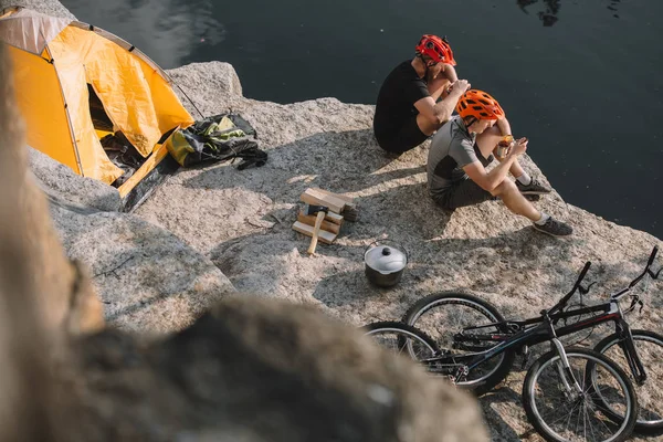 Visão de alto ângulo de viajantes de bicicleta ativos comendo alimentos enlatados no acampamento em penhasco rochoso — Fotografia de Stock