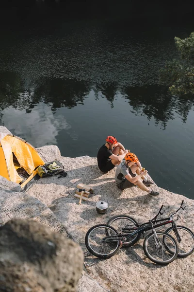 Visão de alto ângulo de jovens viajantes de bicicleta comendo comida enlatada no acampamento em penhasco rochoso — Fotografia de Stock