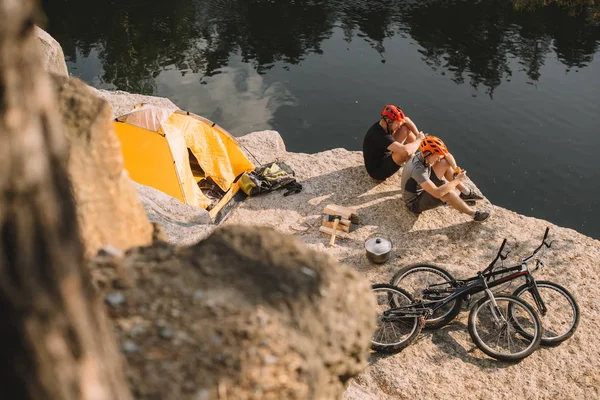 Vista de ángulo alto de ciclistas de prueba activos comiendo comida enlatada en acampada en acantilado rocoso — Stock Photo
