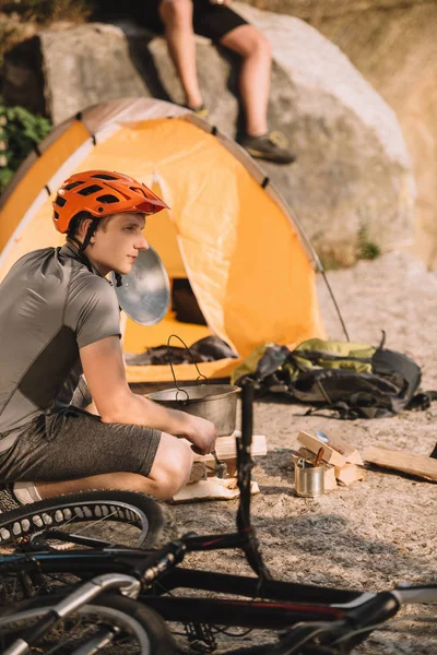 Atlético jovem bicicleta viajante sentado em rochas com barraca de acampamento e amigo borrado sentado no fundo — Fotografia de Stock