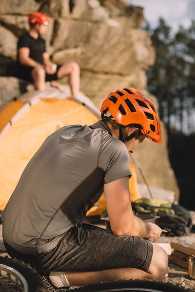 Jeune voyageur à vélo assis sur des rochers avec tente de camping et ami flou assis sur le fond — Photo de stock