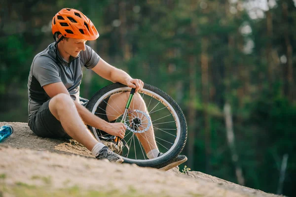 Young trial biker pumping wheel of bicycle outdoors while sitting on stone — Stock Photo