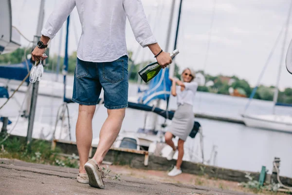 Recortado tiro de hombre sosteniendo copas de vino y botella mientras chica feliz de pie cerca del yate - foto de stock