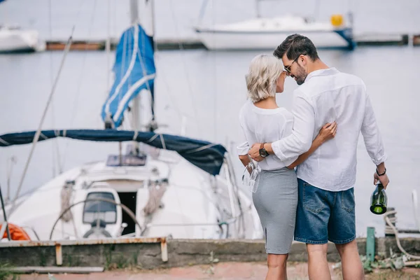 Happy young couple with wine bottle and glasses hugging while standing near yacht — Stock Photo