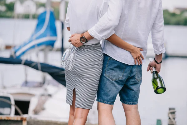 Plan recadré de jeune couple tenant verres à vin et bouteille, étreignant au bord de la mer — Photo de stock