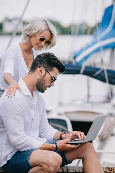 Vue latérale de fille souriante regardant bel homme dans des lunettes de soleil en utilisant un ordinateur portable sur le yacht — Photo de stock