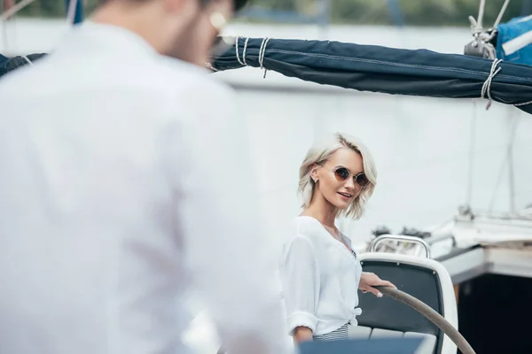 Селективное внимание красивой улыбающейся девушки в солнечных очках, смотрящей на мужчину на яхте — стоковое фото