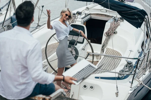 High angle view of smiling girl at steering wheel waving hand and looking at young man on yacht — Stock Photo