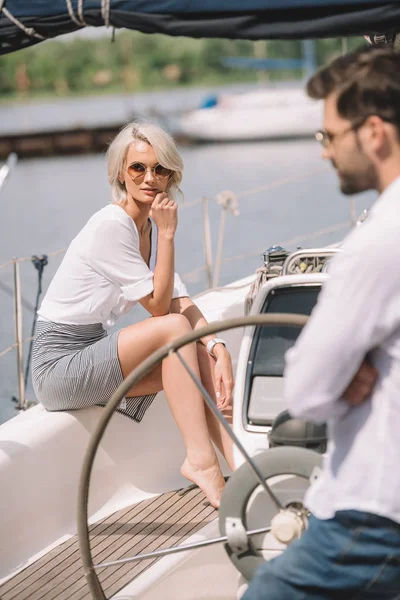 Beautiful pensive girl in sunglasses sitting on yacht and man standing on foreground — Stock Photo