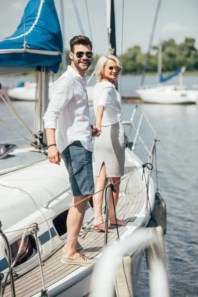 Beau jeune couple heureux dans des lunettes de soleil tenant la main et souriant à la caméra sur yacht — Photo de stock