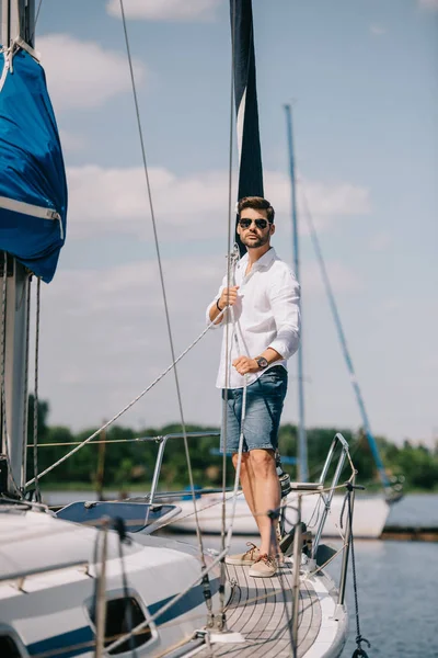 Vue pleine longueur de beau jeune homme en lunettes de soleil debout sur le yacht — Photo de stock