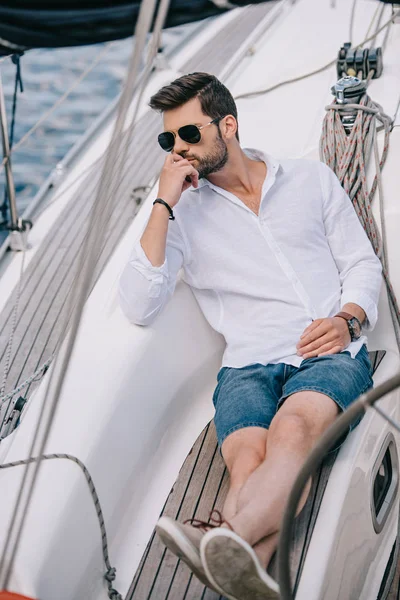Высокий угол зрения застенчивого молодого человека в солнцезащитных очках, который смотрит в сторону, сидя на яхте — стоковое фото