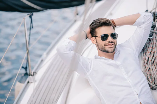 Guapo joven sonriente en gafas de sol descansando con las manos detrás de la cabeza en el yate — Stock Photo