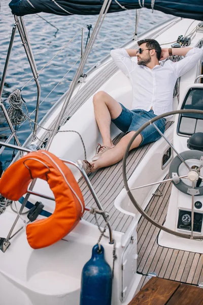 Високий кут зору красивий молодий чоловік в сонцезахисних окулярах дивиться далеко, відпочиваючи на яхті — стокове фото