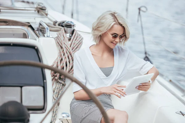 Smiling blonde girl in sunglasses using digital tablet while sitting on yacht — Stock Photo