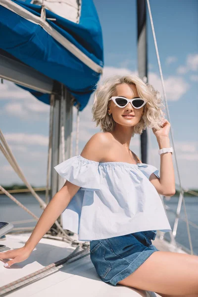 Schöne junge blonde Frau mit Sonnenbrille lächelt in die Kamera, während sie auf einer Jacht sitzt — Stockfoto