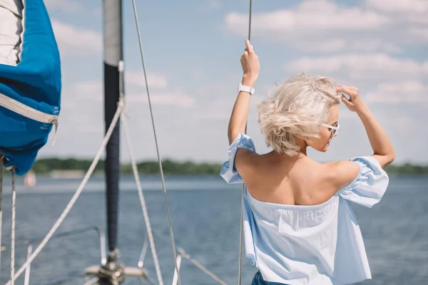 Vue arrière de belle jeune femme blonde en lunettes de soleil regardant loin tout en se tenant debout sur le yacht — Photo de stock