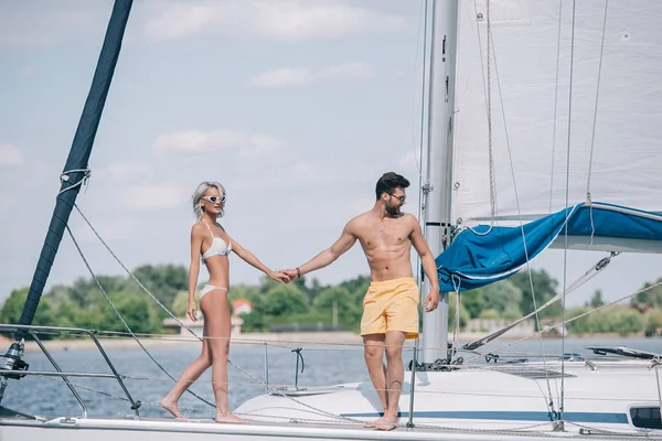Красивая молодая пара в купальниках и солнцезащитных очках держатся за руки, проводя время вместе на яхте — стоковое фото