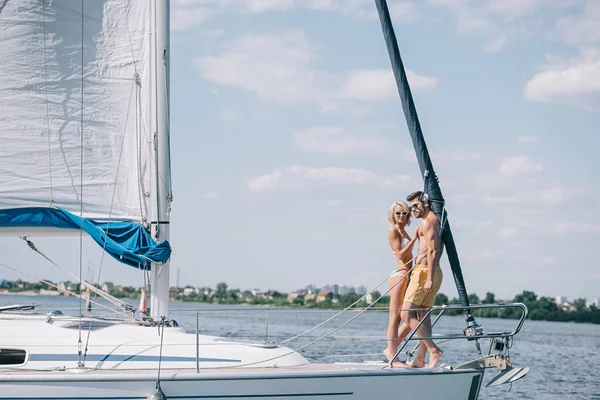 Vue pleine longueur de beau jeune couple en maillots de bain et lunettes de soleil debout ensemble sur le yacht — Photo de stock