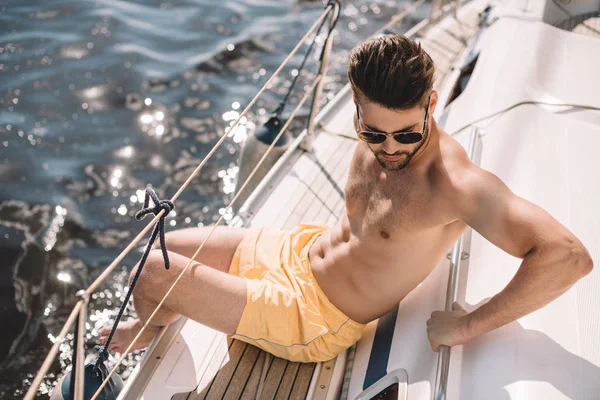Shirtless muscular man in swim trunks and sunglasses having sunbath on yacht — Stock Photo