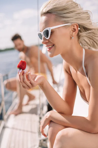 Selektiver Fokus einer lächelnden Frau mit Sonnenbrille, die Erdbeere isst, und ihres Freundes, der hinten auf einer Jacht sitzt — Stockfoto