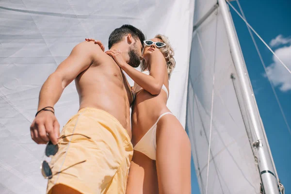 Low angle view of smiling shirtless man embracing attractive girlfriend in bikini on yacht — Stock Photo