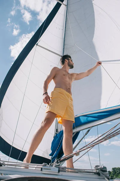 Niedrige Winkelaufnahme des hemdslosen, muskulösen Mannes in Badehose, der das Segel auf der Jacht justiert — Stockfoto