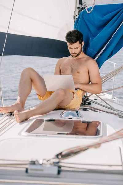 Hombre sin camisa enfocado en bañador utilizando el ordenador portátil en el yate - foto de stock