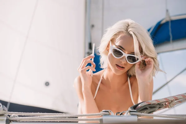 Atractiva mujer en gafas de sol hablando en el teléfono inteligente y la puesta en yate - foto de stock