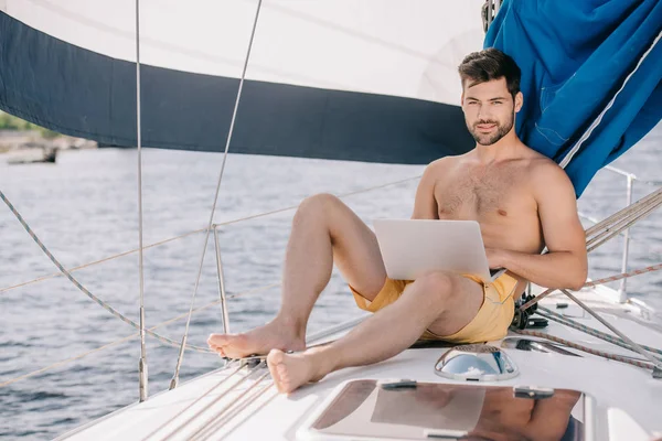 Handsome shirtless man in swim trunks using laptop on yacht — Stock Photo
