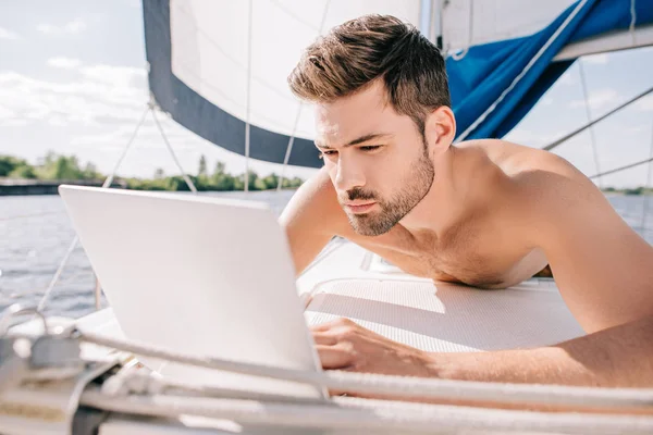 Young shirtless man having sunbath and using laptop on yacht — Stock Photo