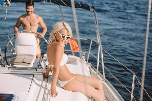 Schöne junge Frau im Bikini beim Sonnenbad, während ihr Freund die Jacht steuert — Stockfoto
