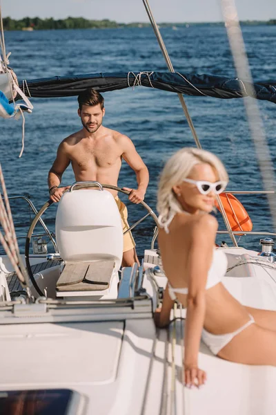 Atractiva mujer joven en bikini tomando el sol mientras su novio yate de dirección - foto de stock