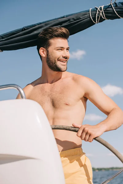 Усміхнений без сорочки м'язистий чоловік у плавальних стовбурах керуючої яхти — стокове фото