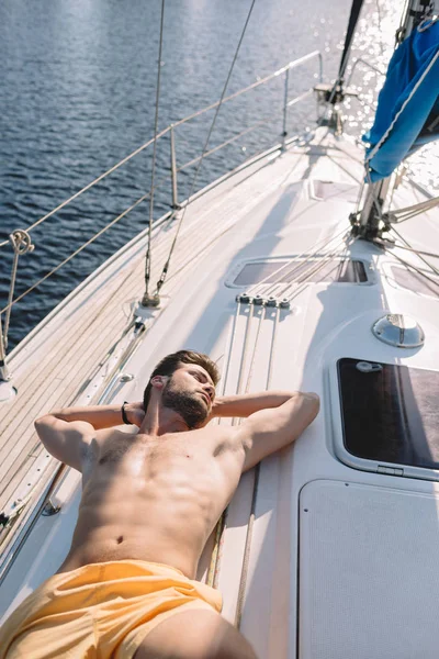 Високий кут зору без сорочки м'язистий чоловік у плавальних стовбурах, що мають сонячну ванну на яхті — стокове фото
