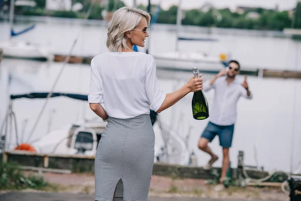 Selective focus of young woman with bottle of champagne and her boyfriend behind on blurred background — Stock Photo