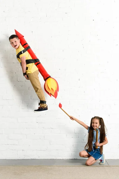 Feliz hermana pretendiendo poner en fuego cohete de juguete con el hermano en casa - foto de stock