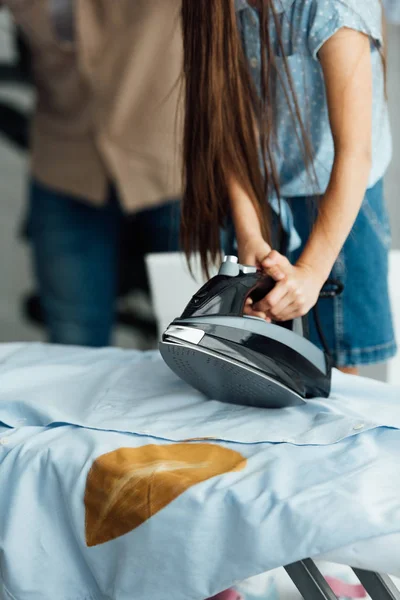 Imagem cortada de filha segurando ferro acima da camisa queimada em casa — Fotografia de Stock