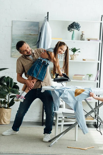 Сердитий батько, що носить неслухняну дочку, і вона хоче прасувати одяг вдома — стокове фото