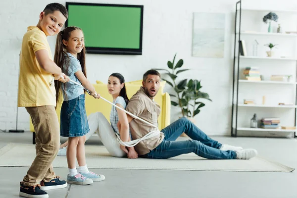 Happy kids tying parents with rope on floor at home — Stock Photo