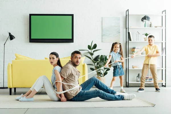 Smiling children tying parents with rope on floor at home — Stock Photo
