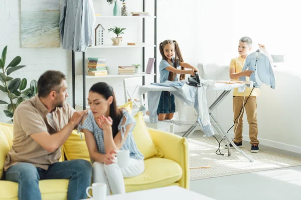 Children ironing clothes and father hugging mother at home, parenthood concept — Stock Photo