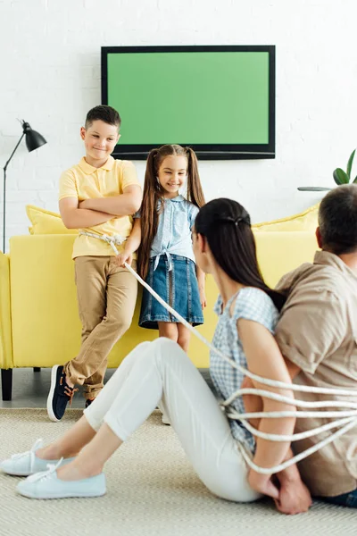 Батьки сидять пов'язані з мотузкою на підлозі і усміхнені діти дивляться на них — стокове фото