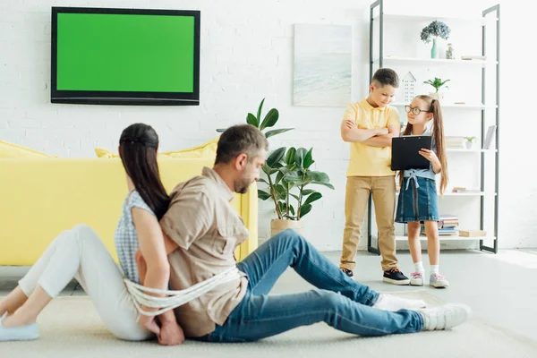 Parents sitting tied with rope on floor and children looking at clipboard at home — Stock Photo