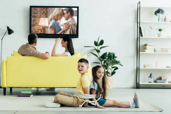 Родители смотрят кино и дети сидят привязанные веревкой на полу дома — стоковое фото