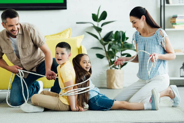 Genitori sorridenti legare i bambini con la corda a casa, concetto di genitorialità — Foto stock