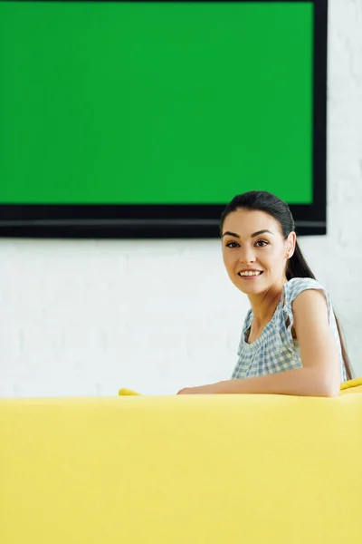 Attraente donna seduta sul divano giallo a casa e guardando la fotocamera — Foto stock