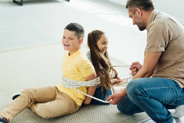 Father tying smiling children with rope at home — Stock Photo