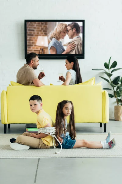 Genitori a riposo con champagne e bambini seduti legati con corda sul pavimento a casa — Foto stock